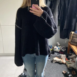 Svart oversized stickad tröja - Säljer en svart oversized stickad tröja med vita dekorativa sömmar längs ärmar och axlar. Tröjan har långa, vida ärmar och en avslappnad passform som ger en cool och chill vibe. Perfekt att slänga på sig när det är kallt ute.