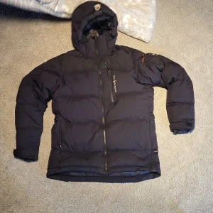 Svart dunjacka Sail Racing M - Säljer en svart dunjacka från Sail Racing, modell Sail Antarctica Expedition. Jackan har huva, dragkedja framtill och bröstficka med logga. Perfekt för kalla dagar med sin tjocka, puffiga design och snygga detaljer.