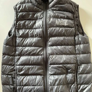 Grå dunväst från Emporio Armani - Snygg grå dunväst från Emporio Armani med quiltad design och logotyp på bröstet. Västen har dragkedja framtill och är tillverkad i polyester. Perfekt att slänga över en hoodie för extra värme. Stilren och sportig look.