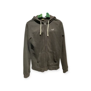 Svart hoodie från Hollister med dragkedja - Svart hoodie från Hollister med vit logga på bröstet, huva med snörning och dragkedja framtill. Mjuk bomullskänsla och två fickor framtill. Perfekt för chill dagar eller när du vill ha en enkel och stilren look.