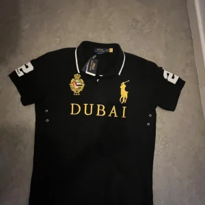 Svart pikétröja Dubai Polo Ralph Lauren - Svart pikétröja från Polo Ralph Lauren med Dubai-brodyr i gult på bröstet, gul polospelare och UAE-emblem. Vit nummer 2 på ryggen och ärmen, samt vit kant på kragen. Klassisk passform och mjukt bomullsmaterial. Perfekt för dig som gillar sportig och exklusiv stil.