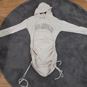 Hoodie-klänning med dragsko "Los Angeles" - En trendig och bekväm hoodie-klänning i en ljus, neutral färg. Klänningen har ett tryck på framsidan med texten "LOS ANGELES CALIFORNIA". Längs sidorna finns justerbara dragskor som skapar en smickrande, rynkad effekt och gör att du kan variera längden. Klänningen har även huva och långa ärmar, vilket gör den perfekt för en cool och avslappnad stil