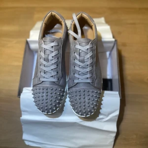 Grå Louboutin sneakers med nitar - Säljer ett par grå sneakers från Christian Louboutin med coola nitar över tån och sidorna. Skorna har snörning och en ikonisk röd sula. Tillverkade i mocka med beige insida, levereras med originalkartong och dustbag. Perfekt för dig som vill sticka ut.