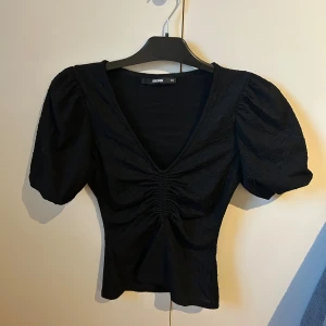 Svart topp med puffärm från BikBok - Snygg svart topp från BikBok i storlek XS med puffiga korta ärmar och v-ringning. Toppen har rynkade detaljer framtill som ger en cool form och sitter figurnära. Perfekt att styla med jeans eller kjol för en trendig look. Tröjan är använd en gång så som ny!