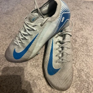 Nike Air Zoom fotbollsskor grå/blå - Säljer ett par Nike Air Zoom fotbollsskor i grått med blå detaljer och stora blå swoosh-loggor. Skorna har snörning, platt sula med dobbar och är tillverkade i syntetmaterial för lätt känsla på planen. Snyggt mönster på hälen och modern, smal passform.