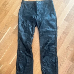 Svarta croco-byxor från GANNI - Säljer ett par svarta byxor från carin Wester croco-imitation. Byxorna har rak passform,  och är tillverkade i ett skinnliknande material med präglat mönster. Stängs med knapp och dragkedja framtill. Perfekta för dig som vill sticka ut.
