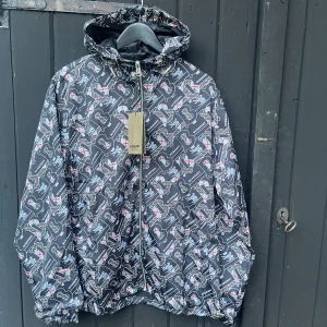 Burberry vindjacka med logomönster. - Snygg svart vindjacka från Burberry i storlek XL med coolt all-over logomönster i grått, vitt och rött. Jackan har huva, hel dragkedja och elastisk nederkant. Perfekt för dig som vill sticka ut med ett lyxigt streetwear-plagg.