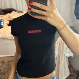 Svart croppad topp Calvin Klein Jeans - Svart croppad topp från Calvin Klein Jeans med korta ärmar och hög hals. Framtill finns en rosa logga. T-shirten är i mjukt bomullsmaterial och har en slim passform som sitter snyggt till jeans eller kjol.