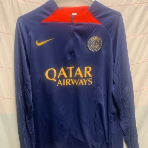 PSG långärmad träningströja Nike M - Säljer en blå långärmad Paris Saint-Germain träningströja från Nike med gul logga och Qatar Airways-tryck. Tröjan har dragkedja vid halsen och detaljer i rött upptill. Materialet är lätt och andas, perfekt för fotbollsträning.