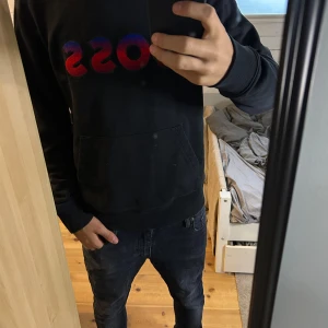 Svart hoodie från BOSS med röd-blå logga - Svart hoodie från BOSS med stor broderad logga i rött och blått på bröstet. Flekarna är bara på spegeln! Sitter bra och passar till massa olika outfits! Pris kan diskuteras! Tveka inte att höra av dig! (Längd=176, vikt=62