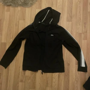 Svart hoodie från BOSS med vita ränder - Svart hoodie från BOSS med dragkedja och huva. Snygga vita ränder längs ärmarna och diskret BOSS-logga på bröstet. Perfekt för en sportig och avslappnad stil. Tillverkad i mjukt material som känns skönt mot huden. Storlek M men passar oxå till S