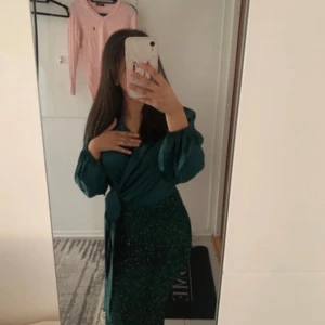 Grön omlottklänning med paljetter - Säljer en elegant grön långklänning med omlottliv och puffiga långärmar. Kjolen är täckt av glittriga paljetter och har en snygg slits bak. Perfekt för dig som vill sticka ut med en glamorös look. Klänningen har en markerad midja och knyt i sidan.