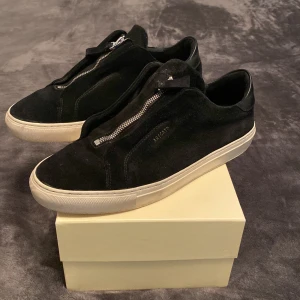 Svarta sneakers från Axel Arigato - Svarta sneakers från Axel Arigato i mocka med dragkedja framtill och vit platt sula. Snygg minimalistisk design med diskret logga på sidan och detaljer i skinn vid hälen. Perfekta för dig som gillar stilrena och moderna skor.