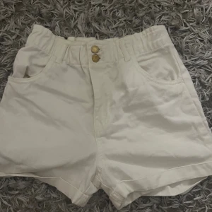 Vita högmidjade shorts från H&M - Snygga vita shorts från H&M med hög midja och två guldknappar framtill. Modellen har bred resår i midjan, framfickor och bakfickor. Materialet är mjuk bomull som känns skönt mot huden. Perfekta för varma dagar och enkel att matcha med din favorit-topp.