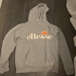Grå Ellesse hoodie med logga - Grå hoodie från Ellesse med stor vit och orange logga framtill. Tröjan har huva med snörning, magficka och ribbade muddar vid ärmslut och nederkant. Perfekt för chill dagar och streetstyle. Det finns tecken på att den är använd som man ser på sista bilden, men för övrigt väldigt fin💞