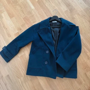 Mörkblå kappa från Design by Si - Snygg mörkblå kappa från Design by Si med klassisk krage och två stora knappar framtill. Jackan har långa ärmar med knappdetalj vid mudden och två rejäla fickor. Säljer då den inte används hos mig 