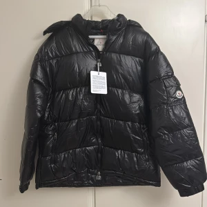Svart Moncler pufferjacka med dragkedja - Svart pufferjacka från Moncler med glansig finish och klassisk logga på ärmen. Är osäker om den är äkta men ser sjukt äkta ut, det står storlek 5xl men folk med storlek 2/3 xl passar den också! Säljer pga för stor. Köpte den för 3200kr. Den är lite skrynklig men måste nog bara hänga till sig! Skriv för fler frågor