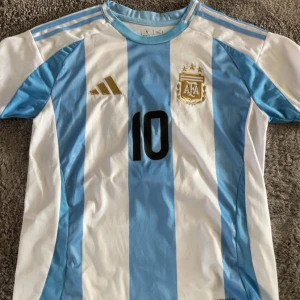 Argentina Messi #10 fotbollströja Adidas - Säljer en officiell Argentina fotbollströja från Adidas med Messi och nummer 10 på ryggen. Tröjan har klassiska ljusblå och vita vertikala ränder, korta ärmar och broderat landslagsmärke på bröstet. Materialet är lätt och andas, perfekt för match eller träning.       Detta är en Replika!