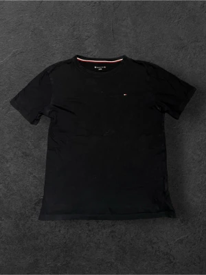 Svart t-shirt från Tommy Hilfiger - Svart t-shirt från Tommy Hilfiger med diskret logga på bröstet och en snygg röd-vit rand på insidan av halsen. Klassisk rund hals och korta ärmar. Tillverkad i mjuk bomull som känns skön mot huden.