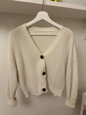 Vit stickad kofta med knappar - Säljer en vit stickad kofta med grov struktur och tre mörka knappar framtill. Koftan har v-ringad hals, långärmade ärmar med uppvik och en bra passform. Perfekt att slänga över en t-shirt för en mysig look. Säljer den då den aldrig har använts 😊