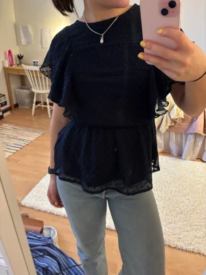 Mörkblå blus med volang och prickar - Säljer en mörkblå blus med volangdetaljer och diskret prickigt mönster. Blusen har korta, luftiga ärmar och markerad midja för en snygg siluett. Tunt, lätt material som ger en flowy känsla. Perfekt att styla med jeans eller kjol.