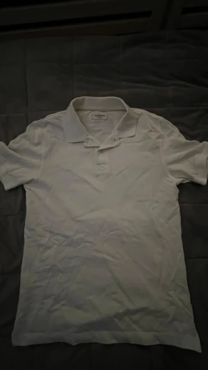 Vit pikétröja från Dressmann S - Klassisk vit pikétröja från Dressmann i storlek S. Tröjan har krage, korta ärmar och knappar framtill. Tillverkad i mjuk bomull som känns skön mot huden. Perfekt för en clean och enkel stil.