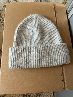 Grå ribbad mössa - Säljer en stilren grå mössa i ribbstickad design. Mössan är tillverkad i en mjuk blandning av ull och polyester, med bred uppvikt kant för extra värme. Perfekt accessoar för kyliga dagar och enkel att matcha med din garderob.