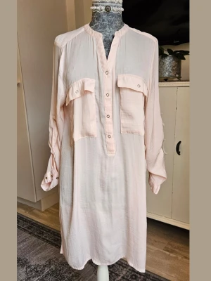 Ljusrosa skjortklänning med fickor - Ljusrosa skjortklänning med långa ärmar och två stora bröstfickor med knappdetaljer. Klänningen har v-ringning med knappar framtill och är gjord i ett tunt, mjukt material som faller snyggt. 