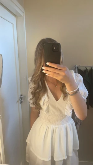 Vit kortklänning med volanger - Säljer en superfin vit kortklänning med v-ringning, puffiga kortärmar och volangdetaljer. Klänningen har smock i midjan och dubbla lager i kjolen som ger en flowy känsla. Perfekt för sommarens alla tillfällen! Materialet känns lätt och luftigt.