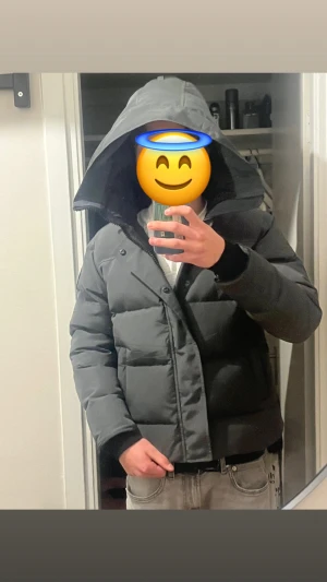 Mörkgrå Canadá Goose pufferjacka med huva - Säljer en stilren mörkgrå pufferjacka med stor huva och svarta detaljer. Jackan har en quiltad design, dragkedja och knappar framtill. Perfekt för kalla dagar och har en loose passform som ger en chill vibe.storlek L fusion,passar Storlek M oxå