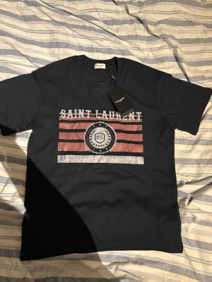 Mörkblå Saint Laurent t-shirt med tryck - Mörkblå t-shirt från Saint Laurent med coolt tryck framtill i rött, vitt och svart. Trycket har ett vintageinspirerat mönster med ränder och emblem. Rund hals och korta ärmar. Materialet är mjuk bomull och passformen är normal.