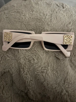 Loewe solglasögon  - Säljer ett par snygga solglasögon från Loewe med rektangulär form och beige bågar. Glasen är svarta och på sidorna finns guldfärgade metalllogos som ger en lyxig touch. Perfekt accessoar för att höja din stil.