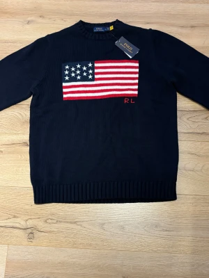 Mörkblå stickad tröja Polo Ralph Lauren - Mörkblå stickad tröja från Polo Ralph Lauren med amerikansk flagga i rött, vitt och blått på bröstet samt RL-broderi. Klassisk rund halsringning och ribbade muddar. Perfekt för dig som gillar preppy stil och vill sticka ut.