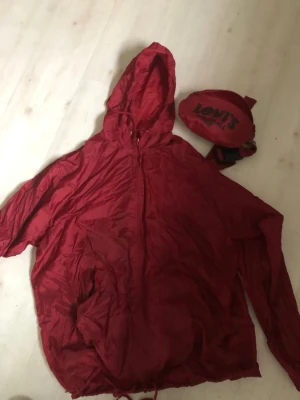 Röd vindjacka från Levi's 501 - Säljer en röd vindjacka från Levi's med huva och dragkedja. Jackan har stor svart 501-print på ryggen och snörning nertill. Till jackan hör en matchande magväska med Levi's-logga. Perfekt för dig som gillar streetstyle och vill sticka ut.