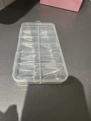 Transparenta lösnaglar i ask - Säljer en praktisk förvaringsask med flera fack fyllda med transparenta lösnaglar i olika storlekar. Naglarna är gjorda av plast och har en snygg spetsig form som passar för både klassisk och trendig nageldesign. Perfekt för dig som gillar att fixa naglarna hemma.