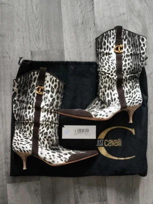 Leopardmönstrade stövletter Just Cavalli - Unika stövletter från Just Cavalli i leopardmönstrad mocka med bruna detaljer och guldfärgad JC-logga. Skorna har spetsig tå och låg klack, vilket ger en cool och edgy look. Perfekt för dig som vill sticka ut med ett statement-plagg. Dustbag ingår, stl 38 sparsamt använda.
