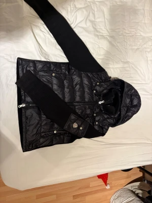 Svart Moncler pufferjacka med huva - Svart pufferjacka från Moncler med stickade ärmar och huva. Jackan har dragkedja framtill, tryckknappar på fickor och Moncler-logga på ärmen. Insidan har ett unikt serietidningsmärke. Perfekt för dig som gillar street och vill ha något som sticker ut. Pris kan diskuteras.