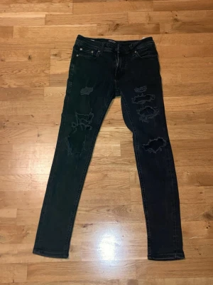 Svarta skinny jeans Jack & Jones 29/30 - Svarta jeans från Jack & Jones med skinny passform och slitna detaljer på benen för en cool look. Klassisk femficksmodell och stretchigt jeanstyg som sitter tajt. Perfekt för dig som gillar streetstyle och vill ha ett par jeans med edge.