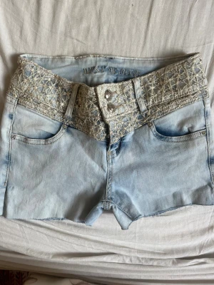  jeansshorts med brodyr - Säljer ett par ljusblå jeansshorts från West Jeans med bred midja och snyggt broderade detaljer i silvertråd både fram och bak. Shortsen har råa kanter och dubbla knappar framtill. Bakfickorna har extra dekorationer med strass. Perfekta för sommaren! 
