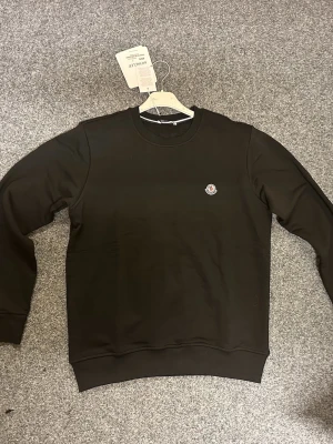 Svart sweatshirt från Moncler - Svart sweatshirt från Moncler med rund hals och diskret logotyp på bröstet. Tröjan har långa ärmar, ribbade muddar och är tillverkad i mjuk bomull. Enkel och stilren design som passar perfekt till jeans eller joggers.
