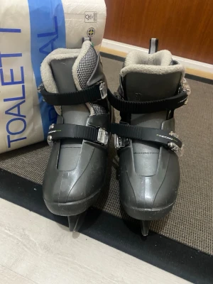 Grå skridskor Tecno Pro Raptor - Säljer ett par grå skridskor från Tecno Pro, modell Raptor. De har två svarta spännen med metallfästen, vit och svart mesh vid skaftet samt mjuk, ljusgrå fodring. Snygga gröna detaljer och logga på sidan. Perfekt för dig som vill glida fram på isen med stil.