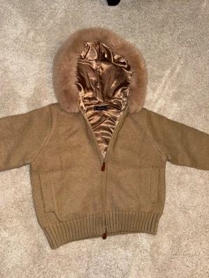 Alessia fur jacket  - Mysig beige stickad jacka från Alessia med huva fodrad i glansigt satin och kantad med fluffig fuskpäls. Jackan har dragkedja framtill, ribbade muddar och två fickor. Perfekt för kyliga dagar när du vill vara både varm och snygg.