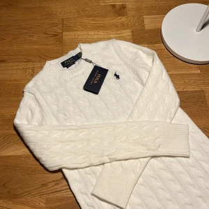 Vit kabelstickad tröja Polo Ralph Lauren - Vit kabelstickad tröja från Polo Ralph Lauren. Klassisk rund halsringning, långa ärmar och broderad marinblå logga på bröstet. Tröjan är gjord i mjuk ullblandning och har ribbade muddar vid ärmslut och nederkant. Aldrig använt den.