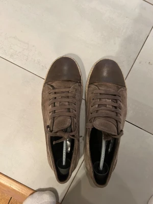 Bruna sneakers från Lanvin i mocka - Snygga bruna sneakers från Lanvin med ovandel i mjuk mocka och tåparti i mörkbrunt skinn. Skorna har rund tå, snörning och en vit platt sula med diskret logga undertill. Perfekt för dig som gillar stilrena och klassiska sneakers med exklusiv känsla. Skorna har små defekter och priset kan diskuteras 
