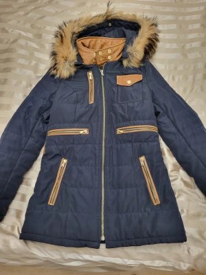 Mörkblå pufferjacka från Ventiuno - Snygg mörkblå pufferjacka från Ventiuno med bruna detaljer i skinnimitation, guldfärgade knappar och dragkedjor. Jackan har en huva med fuskpäls, flera fickor med dragkedja och hög krage med tryckknappar. Perfekt för kalla dagar. Små i storlek,  passar nog M/S