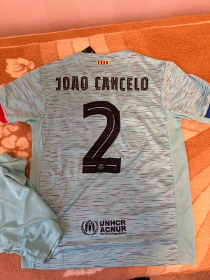 Fc Barcelona third 2023/2024  -  FC Barcelona third matchställ med Joao Cancelo och nummer 2 på ryggen. Tröjan har klubbmärke, Spotify-logga och UNHCR-tryck, samt röda och blå detaljer på axlar och ärmslut. Setet inkluderar matchtröja och shorts i lätt polyester med randigt mönster.