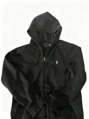 Svart hoodie från Polo Ralph Lauren - Svart hoodie från Polo Ralph Lauren med klassisk broderad logga på bröstet. Tröjan har huva med snörning, dragkedja framtill och långa ärmar. Tillverkad i mjuk bomull som är skön att ha på sig. Perfekt för dig som gillar stilrena och tidlösa plagg.