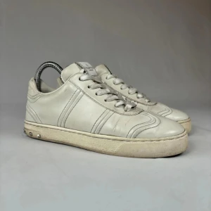 Valentino Garavani vita sneakers - Snygga vita sneakers från Valentino Garavani med klassisk rund tå och snörning. Skorna är tillverkade i skinn och har coola nitar på hälen. Yttersulan har ett unikt kamouflagemönster och logga. Perfekta för dig som gillar stilrena designer med edge.