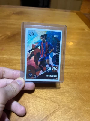 Randig blå-röd fotbollsväst FC Barcelona - Ett officiellt Topps Match Attax-kort med fotbollslegendaren Ronaldinho i Barcelona-tröjan. Kortet tillhör serien Showboat, som lyfter fram spelare kända för teknik och kreativitet. Det har en färgstark, glansig design med Ronaldinho i aktion samt hans värden: 58 DEF och 94 ATT. Kortnumret är 417.