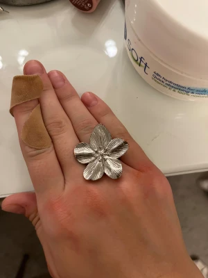 Stor silvrig blomring - Säljer en riktigt snygg och stor ring i silverfärgat metall med en detaljrik blomma som motiv. Blomman har fem blad med tydliga ådringar och ger en cool statement-look. Perfekt för dig som gillar unika accessoarer och vill sticka ut. Köpt för 300kr, ringen är skjusterbar